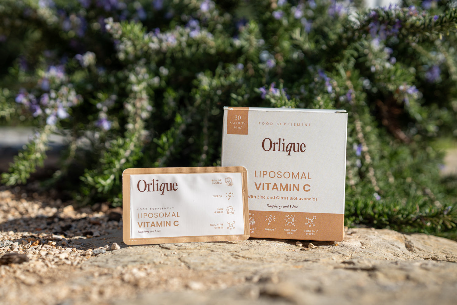 A box of Orique Liposomal Vitamin C with Zinc and Citrus Bioflavonoids is displayed outdoors with floral elements in the background.