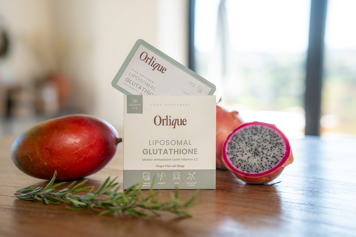A package of Orlique liposomal glutathione supplement is displayed with a mango and a pomegranate in the background.